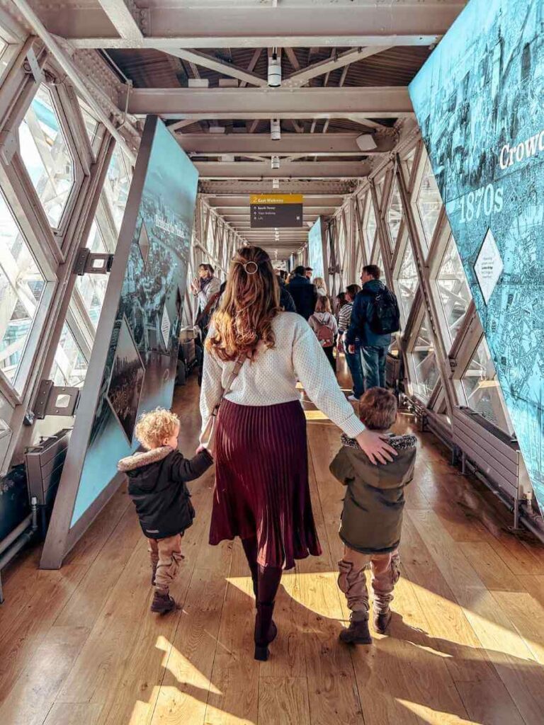 Mum and kids exploring the inside walkway of Tower Bridge with city views behind them
