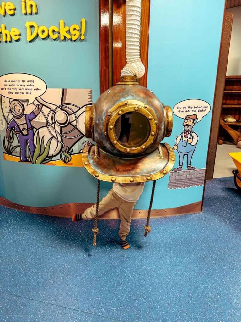 Child trying on a diver’s helmet in the Mudlarks gallery at the Museum of London Docklands, part of the interactive dress-up area.