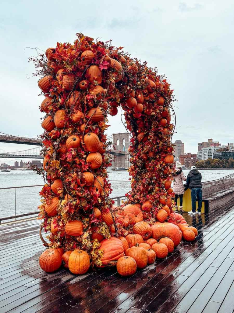 Side view of Pier 17’s iconic pumpkin arch with the Brooklyn Bridge in the background — a autumnal photo spot in NYC in October.