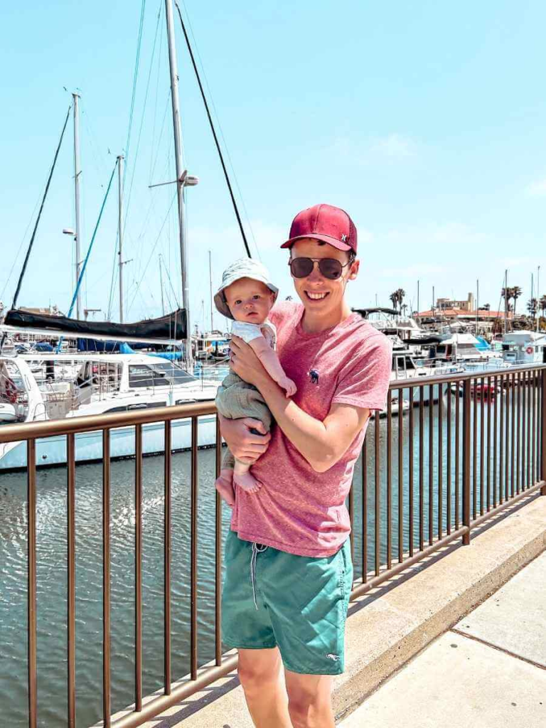 Father and son watching yachts and sailboats docked at Santa Barbara Harbor, with the ocean and marina walkway in view.
