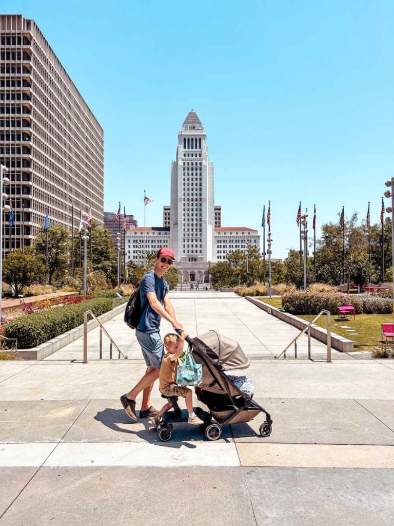 Dad pushing the Joolz Aer+ stroller with a baby seated and toddler riding on the attached footboard.